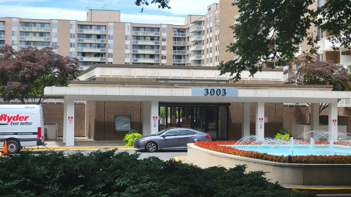 A brown and white brick apartment building with a water fountain and cars parked in front.