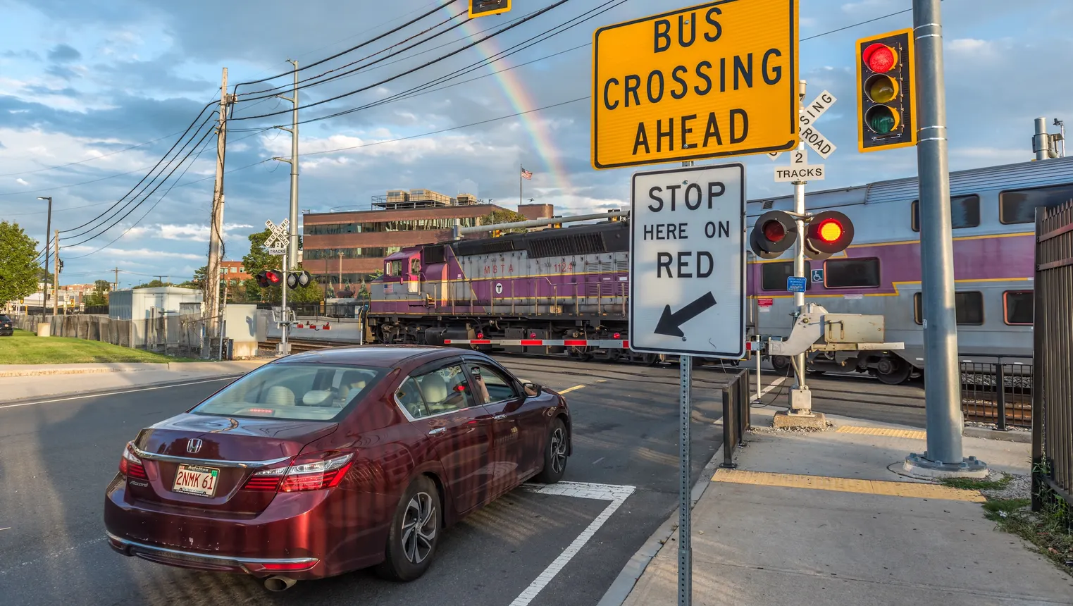 A maroon sedan stopped at a railroad crossing a a purple and silver commuter train passes.