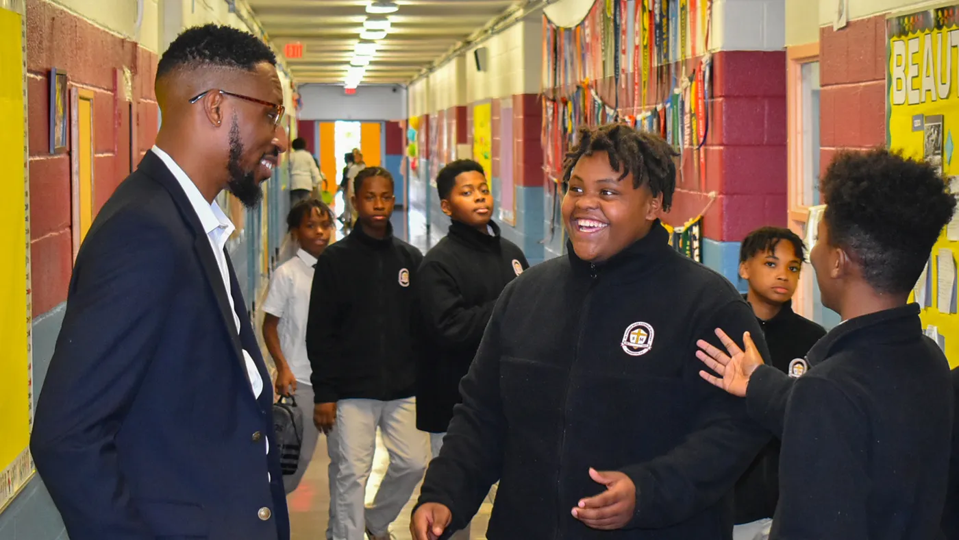 An adults stands in a school hallway talking to a student. Other students are passing by in the hallway