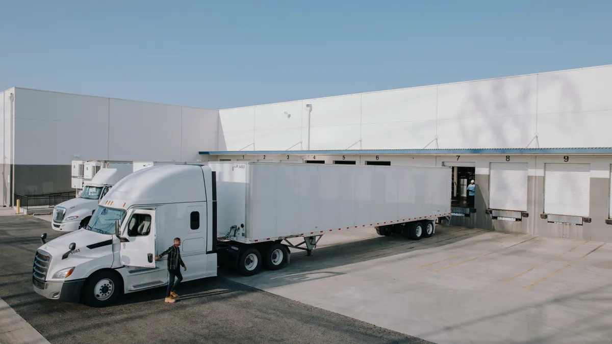 A truck driver exits a tractor-trailer after arriving at a docking area, where a worker can be seeing in a docking bay.