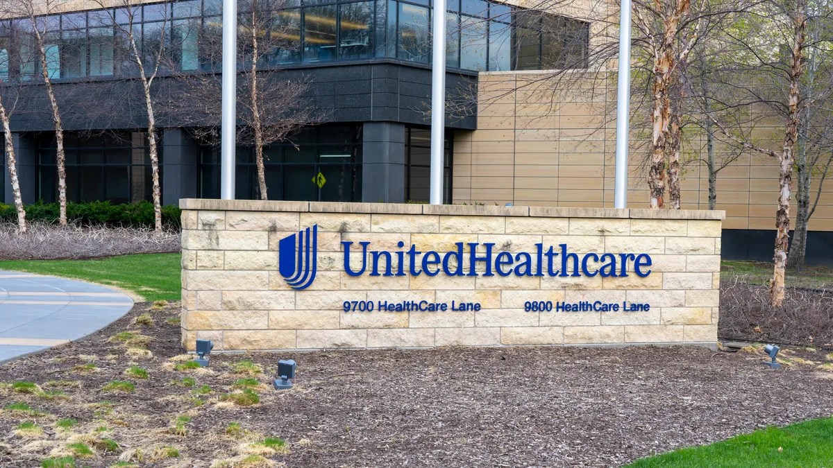 A sign with the words UnitedHealthcare is fixed to a low stone wall outside of an office building.