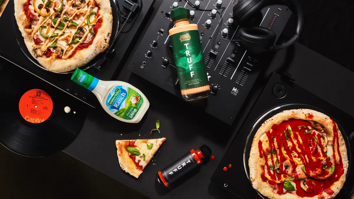 Bottles of Spicy Truffle Ranch, Hidden Valley Ranch and Truff hot sauce lie on an audio mixer. Pizzas sit on two record turntables, and a pair of black headphones and a slice of pizza sit nearby.