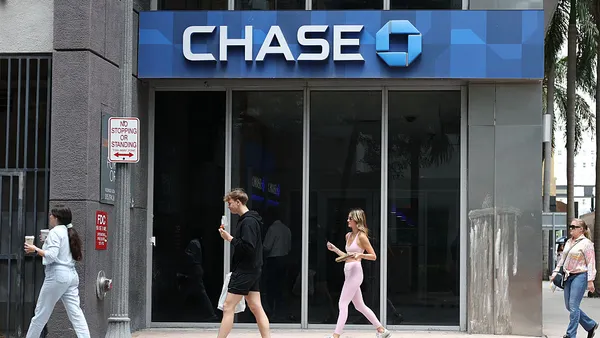 A bank branch building with the word "Chase" and the Chase logo on a blue sign is shown, with people walking past in the foreground.