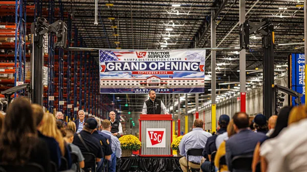 A person speaks in front of a crowd inside a warehouse, with a "Grand Opening" banner in the background.