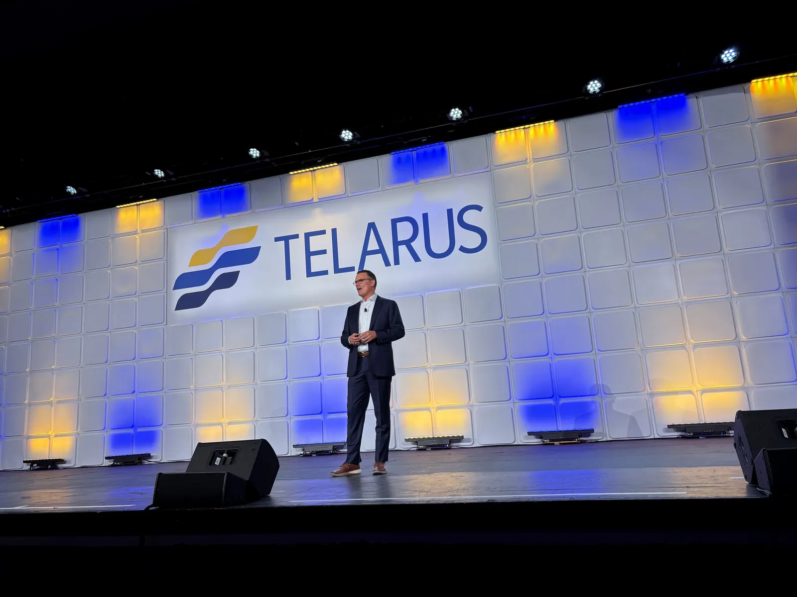 suited man speaking on stage in front of a sign that says telarus