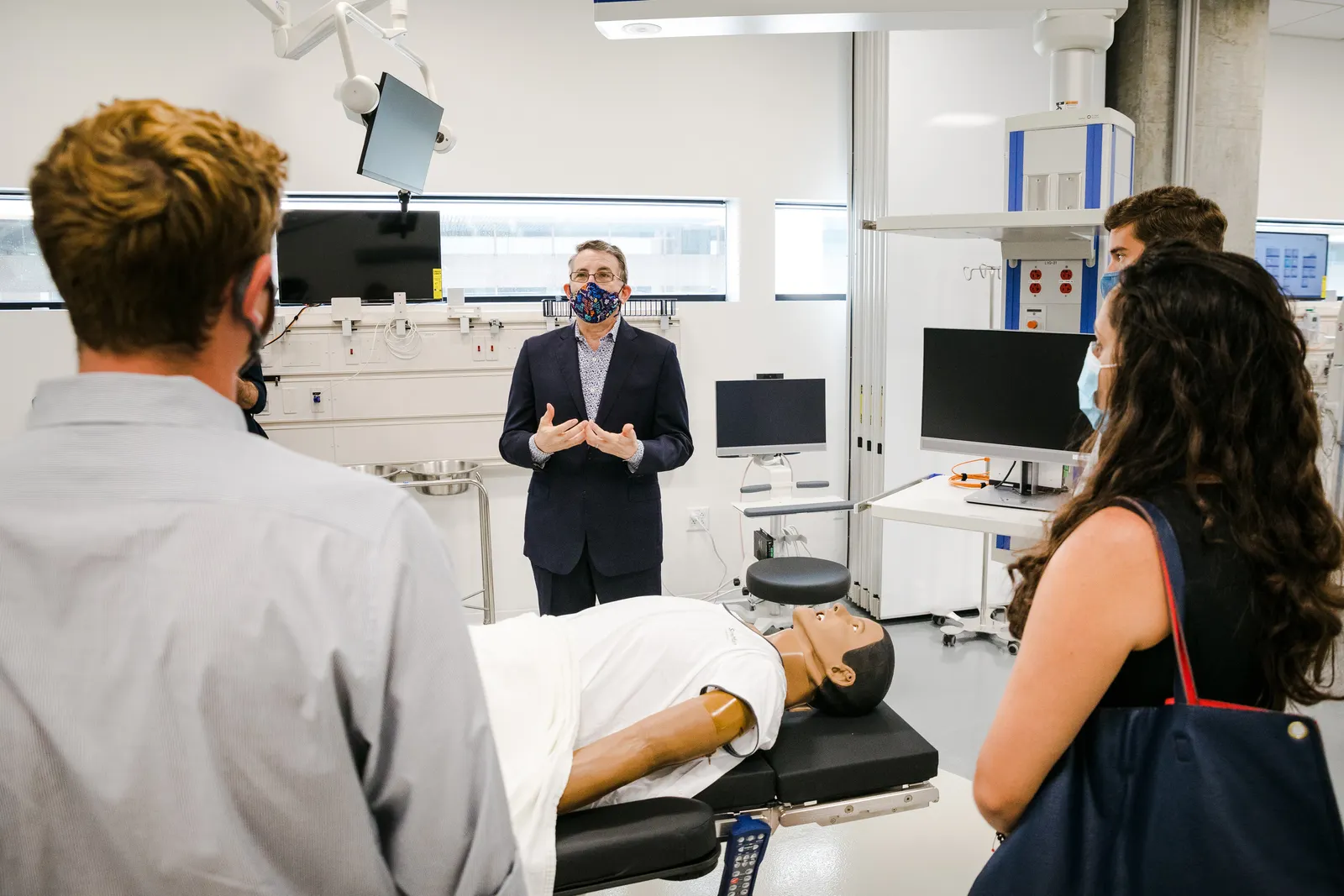 Mark Schuster, wearing a mask, stands in front of a simulation lab. A simulated human body lays on a table while three medical students watch.