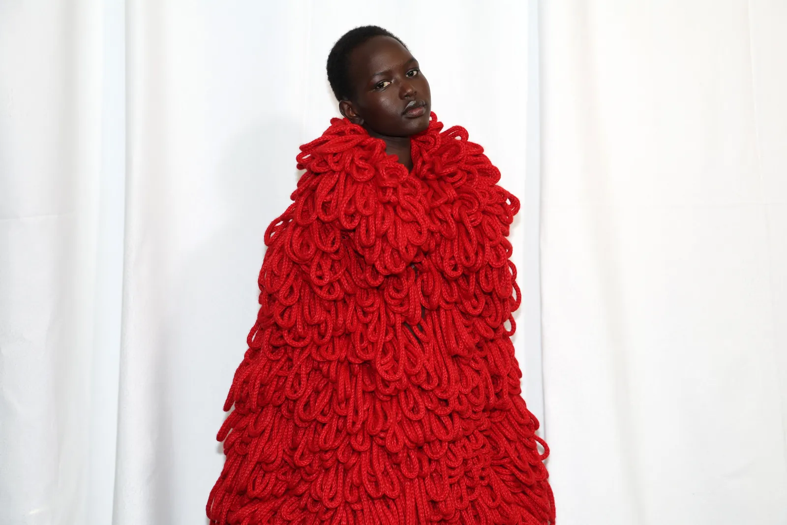 A model wears a large red knit shawl backstage at a fashion show.