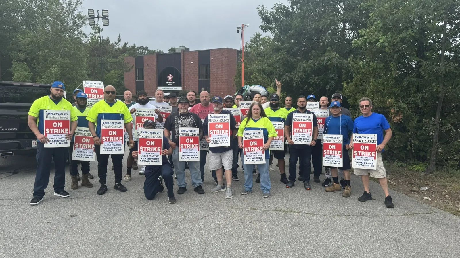 Workers stand in a group outside with signs that read "Employees of Republic Services On strike - Unfair Labor Practice - Teamsters Local No. 25"