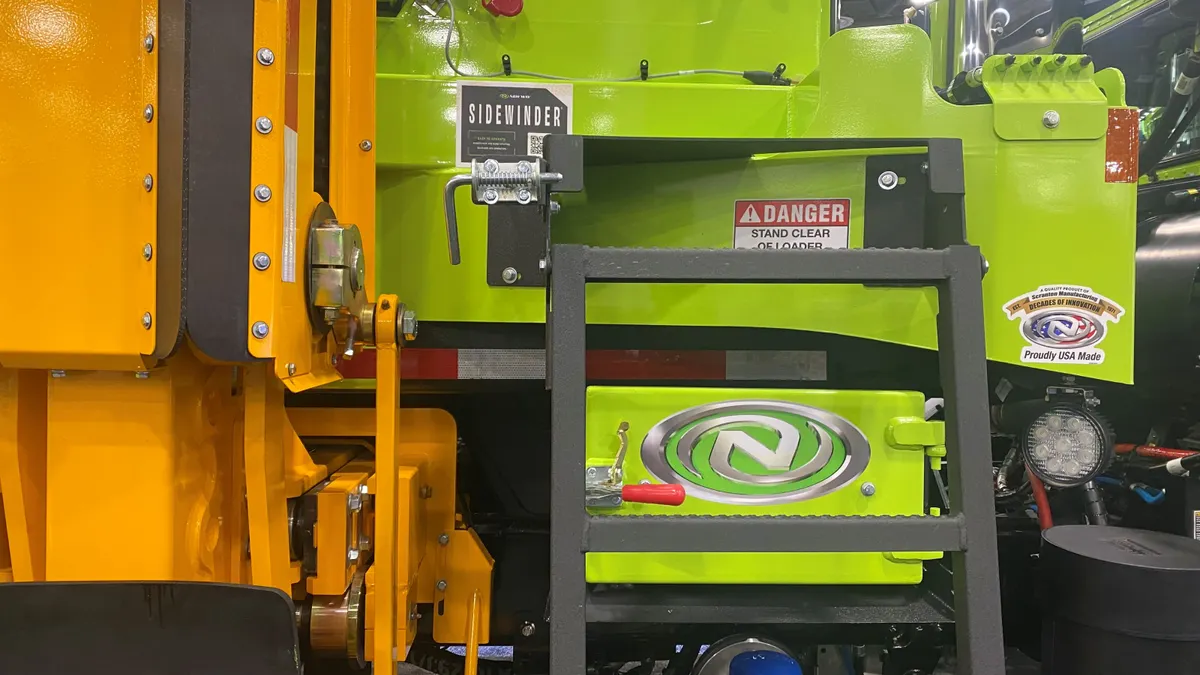 The side of a truck is pictured with multiple logos, including that of New Way Trucks. The company's automated side-loading mechanism is partially in view.