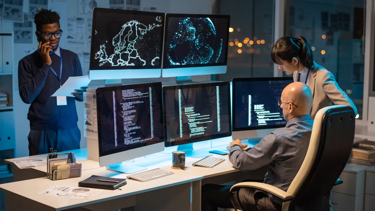 Two people look at five computer screens showing code and digital maps in an office. A third person talks on the phone while looking at a paper.