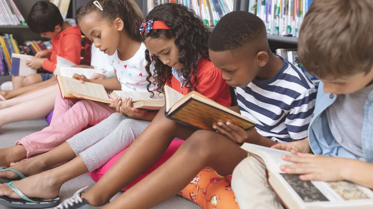 Side view of mixed ethnicity school kids sitting on cushions and studying over books in a library at school against bookshelves in background.