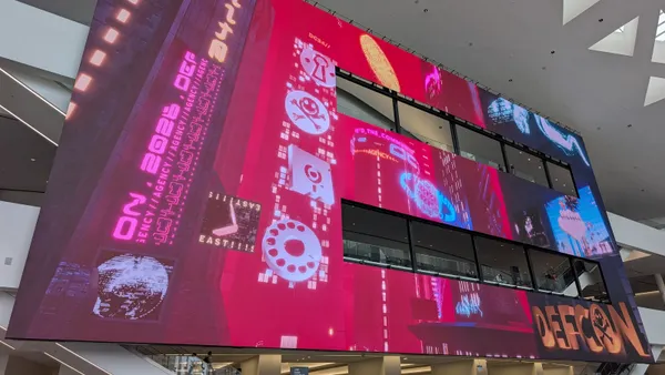 A large screen inside a convention center displays logos and other imagery related to the DEF CON cybersecurity conference