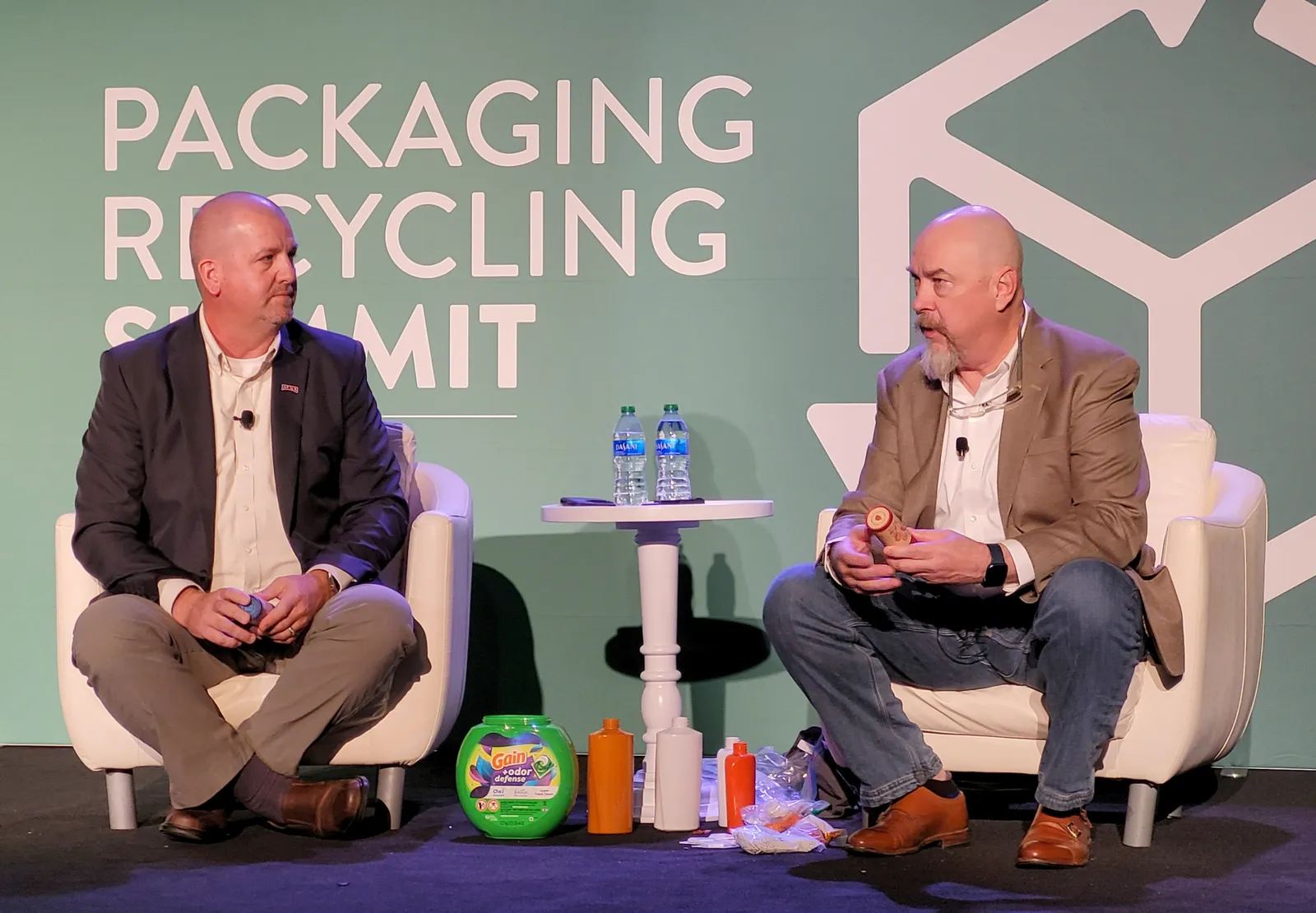 Two people sitting on a stage in front of a green background that says "Packaging Recycling Summit," with various colored product containers between them.