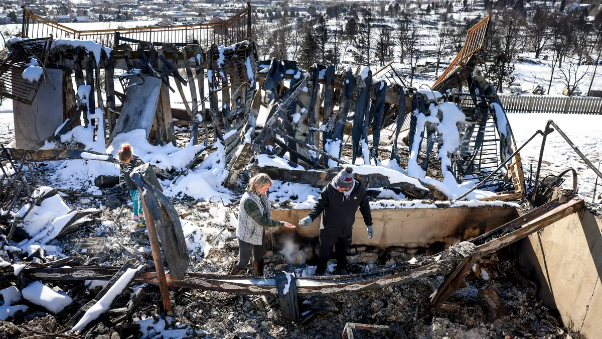 Three people in winter gear walk through the burn-out remains of a building partly coated with snow. Blue mountains are in the background.