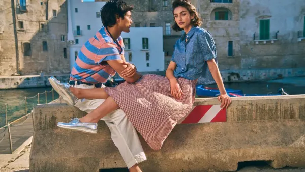 Two people wearing boat shoes and casual clothing sit together on a stone wall outdoors on a sunny day.