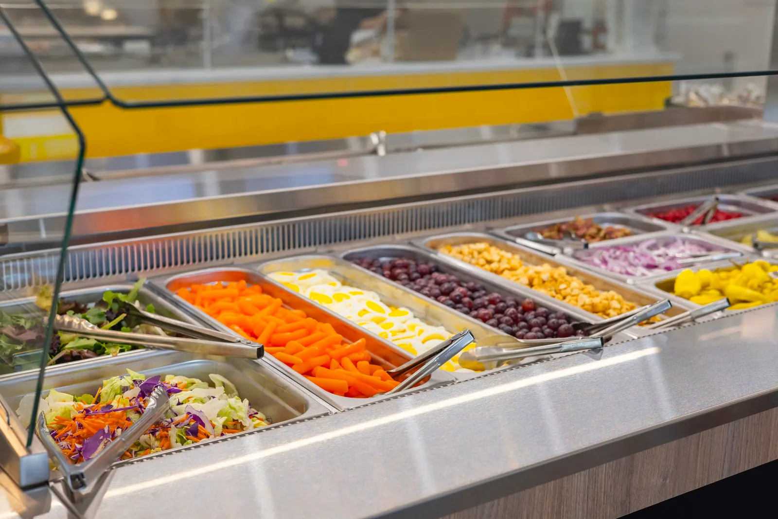 A fresh food bar that includes containers of salad, vegetables, fruit and proteins, each with tongs or a spoon in them for serving.