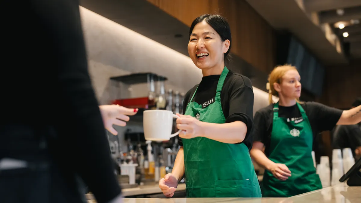 A smiling worker hands someone a ceramic mug of coffee.