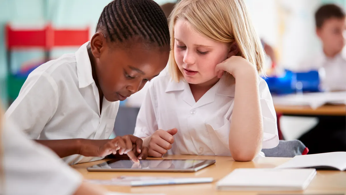 Two students are sitting next to each other at a desk in a classroom and are looking at a tablet