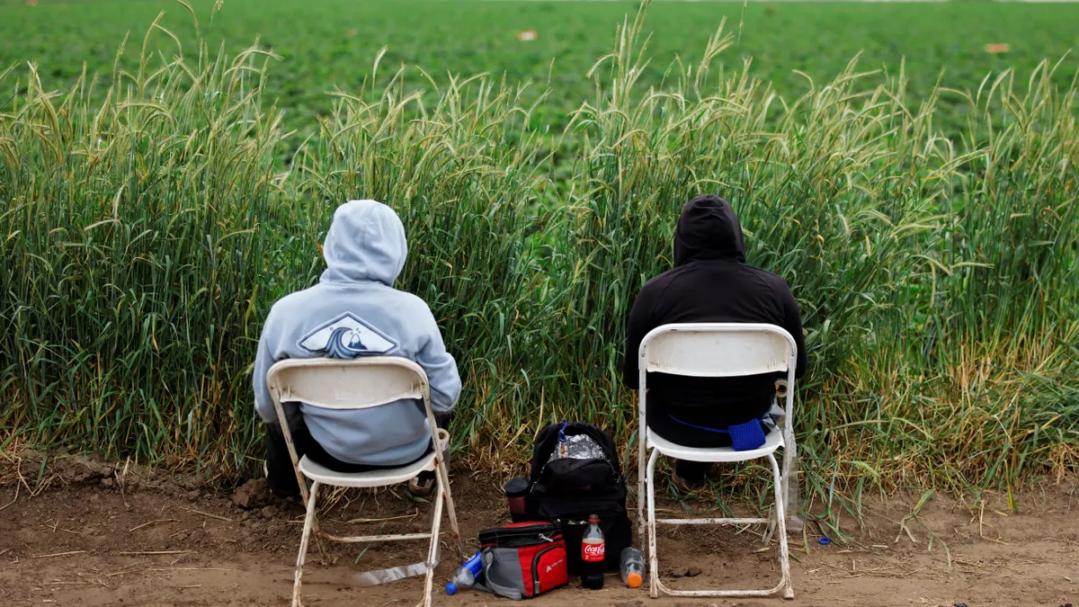 Two individuals sit on folding chairs outdoors, with lunch bags between them.