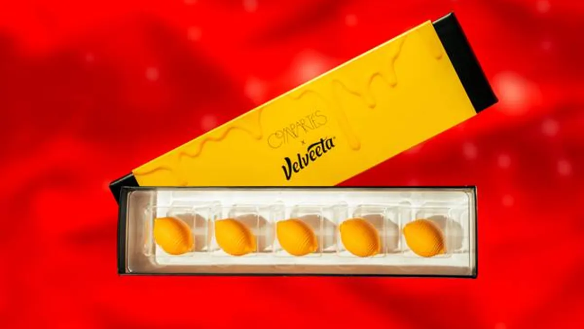 A rectangular box of five TruffVel candies, which look like Velveeta Shells & Cheese, with the orange and black lid behind it. The background is red.