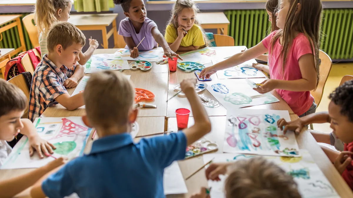 A diverse group of elementary students is seated around a large square table and painting during art class.