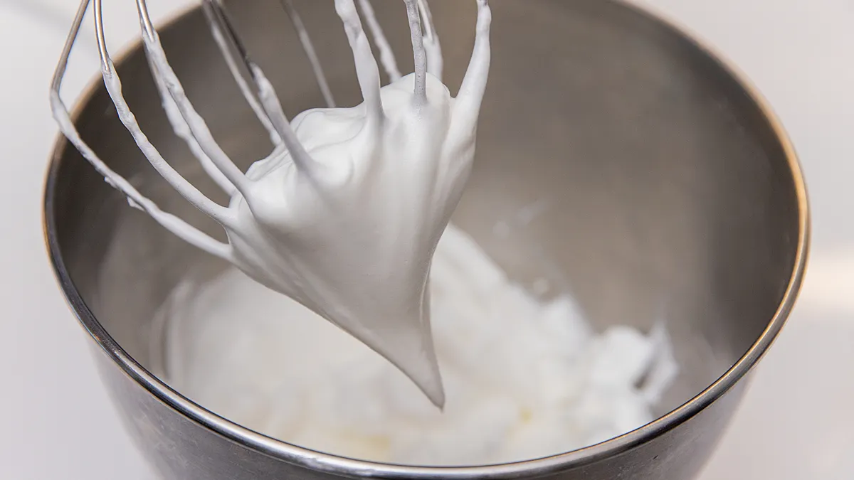 A beater with whipped egg white is above a silver bowl containing more whipped egg white.