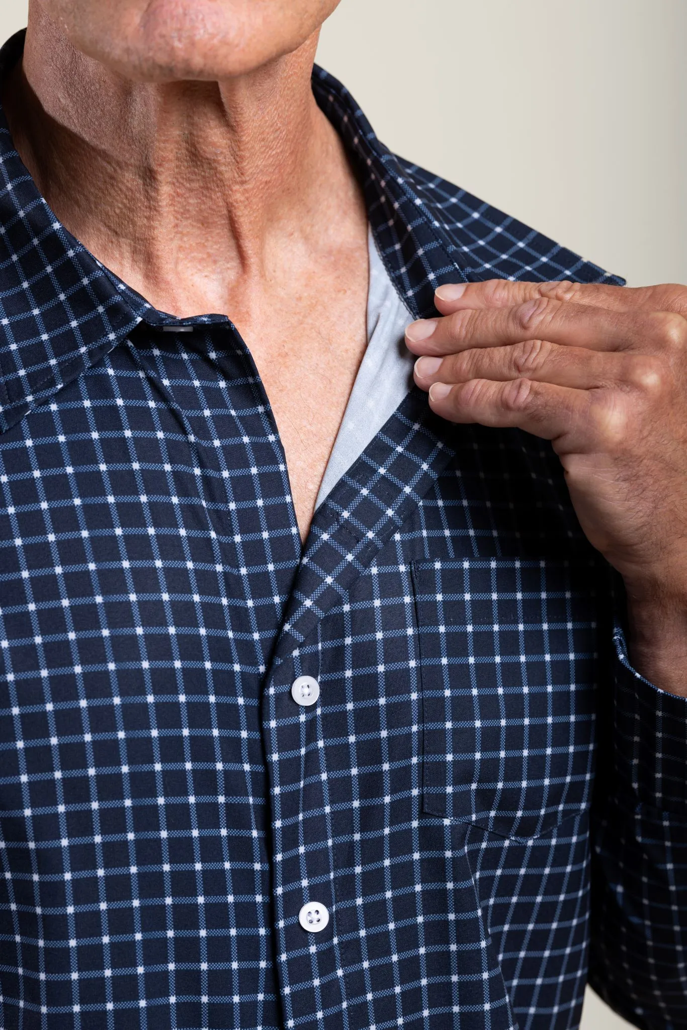 A person wears a navy and white checked long-sleeved shirt with white buttons on the front. A hand pulls the top of the collar back to display the shirt’s magnetic closure.