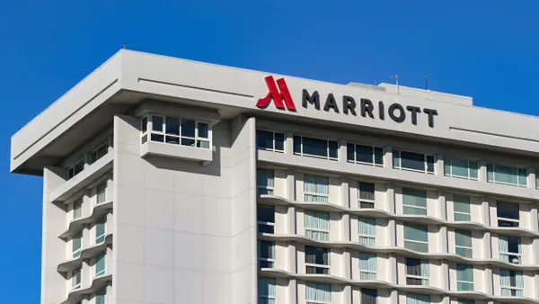 A wide-angle view of a large hotel building with a modern, rectangular design features rows of evenly spaced windows, a white and beige exterior, and the Marriott logo located at the top of the building's roof edge. 