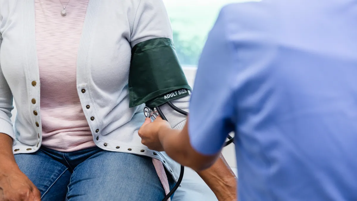 A person in a blue shirt applies a blood pressure cuff to a person wearing a cardigan.