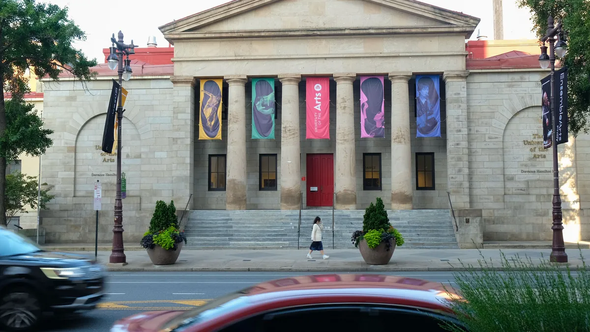 View of building with columns and colored banners as vehicles and a pedestrian pass.