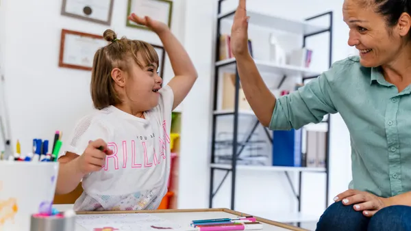 A student and teacher are sitting at a table in a classroom. The student is holding a Crayon and is high fiving the teacher.