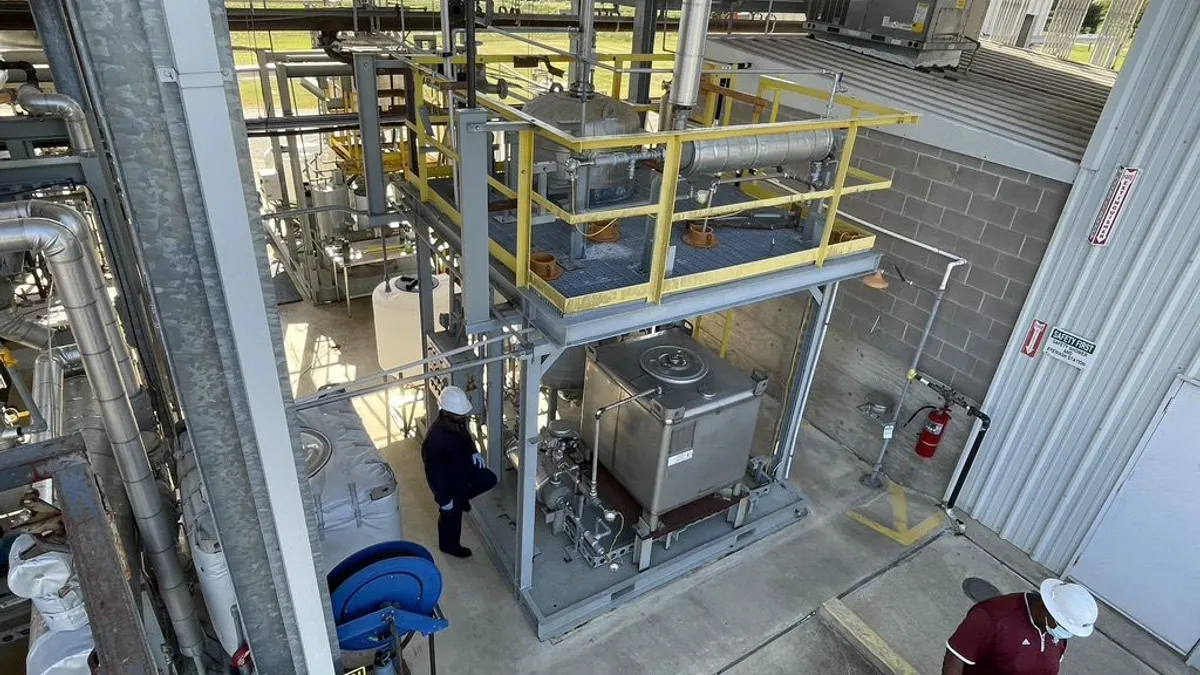 Fermentation equipment in an open building, with concrete block walls and floors on one side, and an open view of grass and trees behind. Two people in hard hats stand nearby.