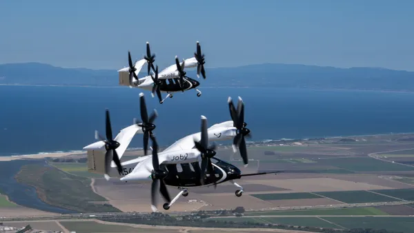 Two futuristic electric vertical takeoff and landing aircraft fly over farmland.
