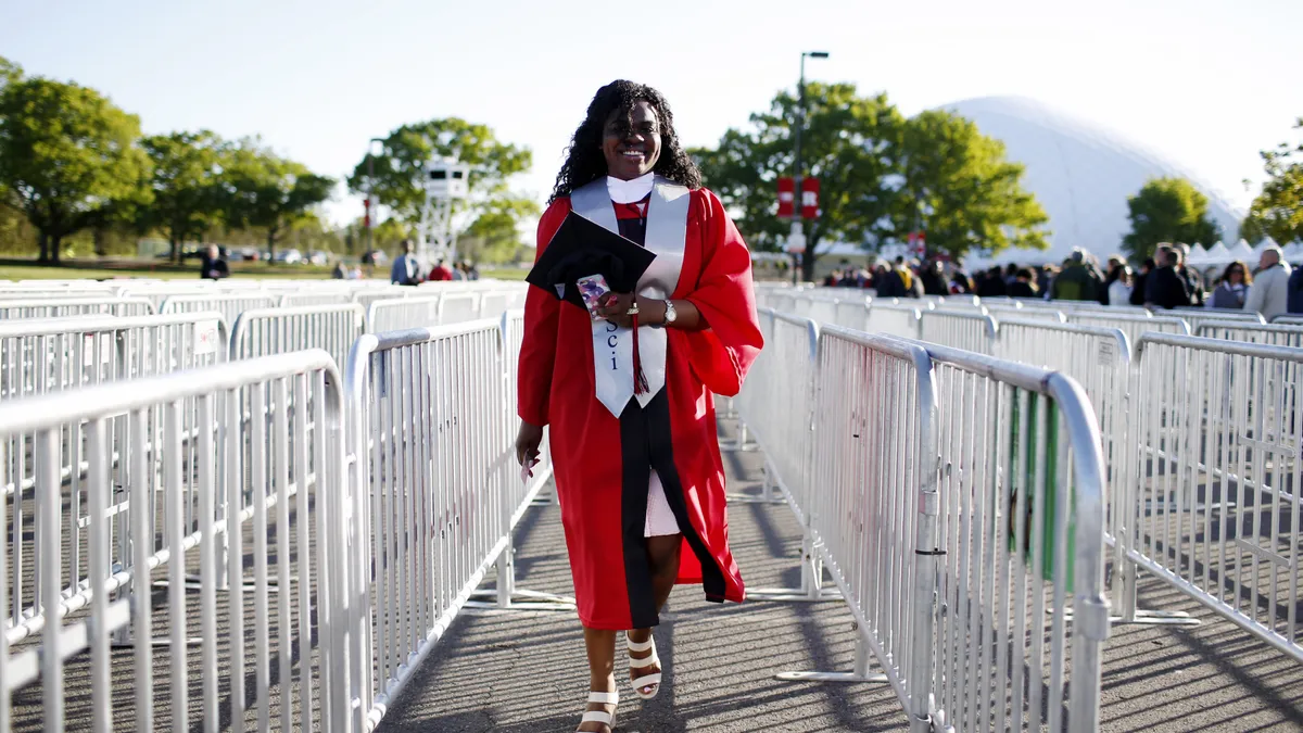 Someone wearing a graduation gown walks through crowd control lines outside.