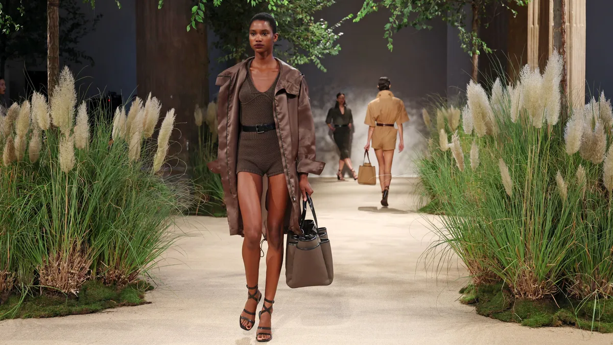 Model in a brown shorts outfit walks on a runway flanked with stalks of wheat.