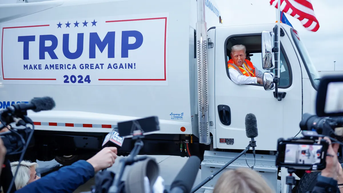 Former President Donald Trump sits in the cab of a garbage truck emblazoned with his campaign's logo as reporters look on.