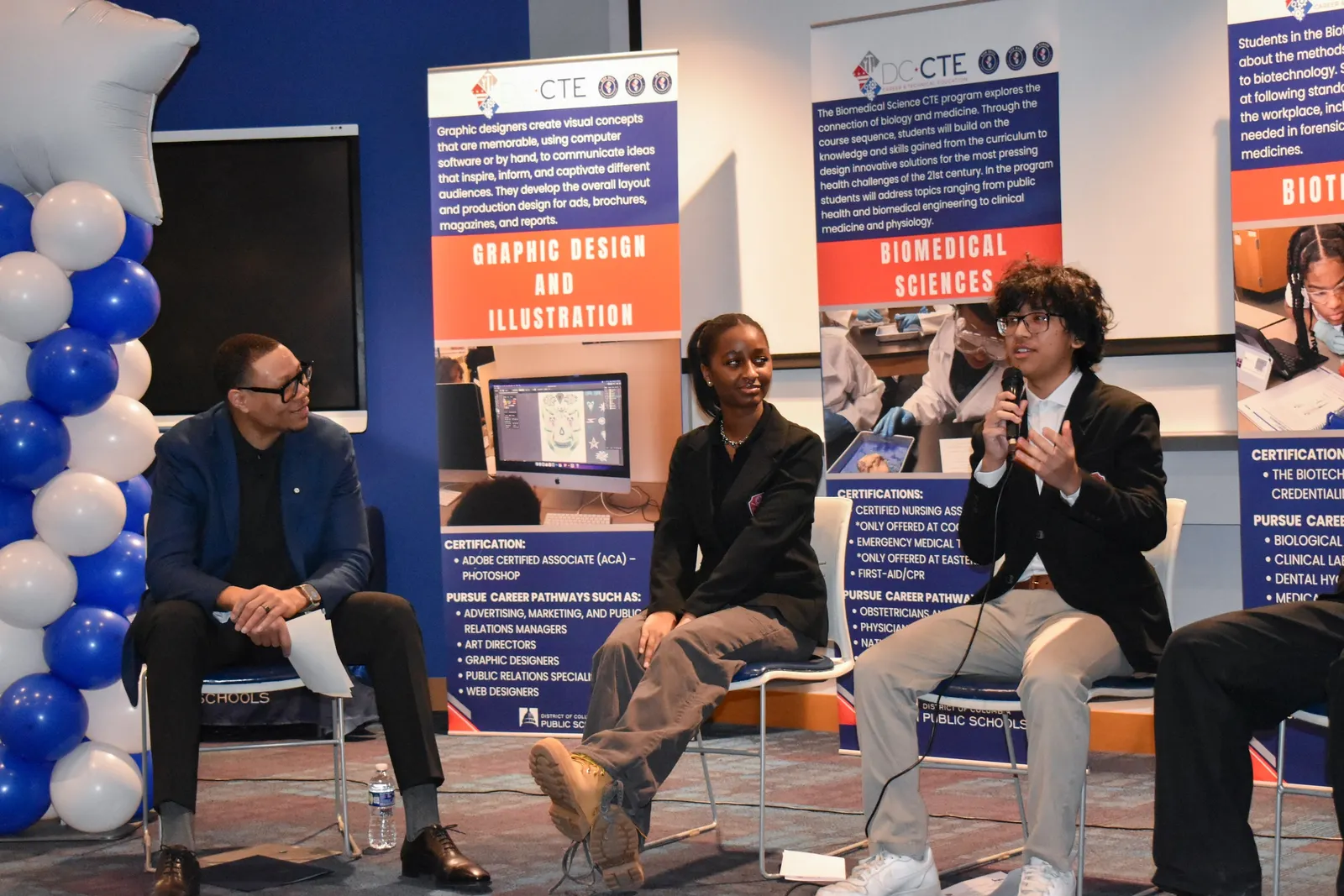 An adult and two students sit in chairs in a room. One student is talking into a microphone. Banners and balloons are near and behind the speakers.