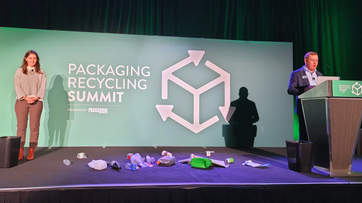 Two people on stage at a recycling event with packaging strewn on the stage and two trash bins.