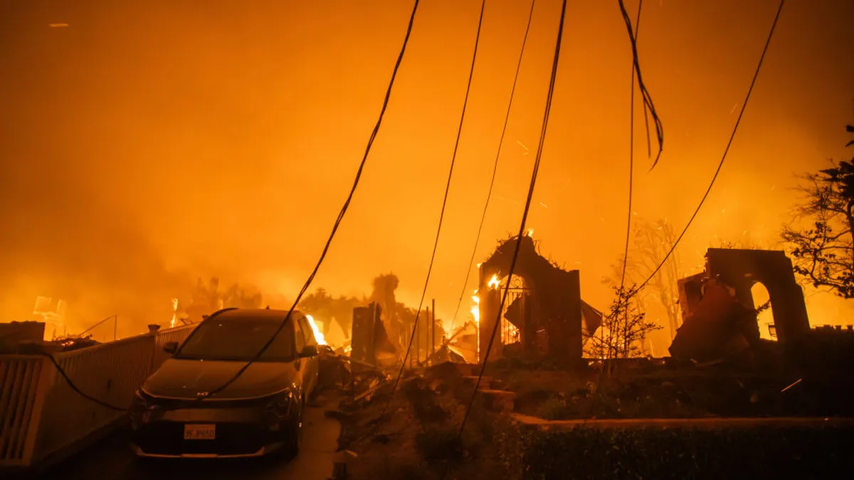 A car, remains of a house and downed power lines stand in front of an orange wall of flame.