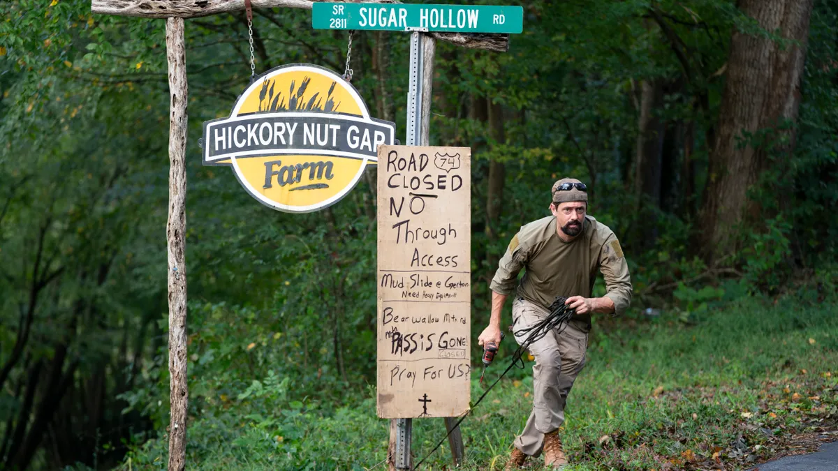 A man is seen putting up a cardboard sign warning of road closures next to a logo for Hickory Nut Gap Farm