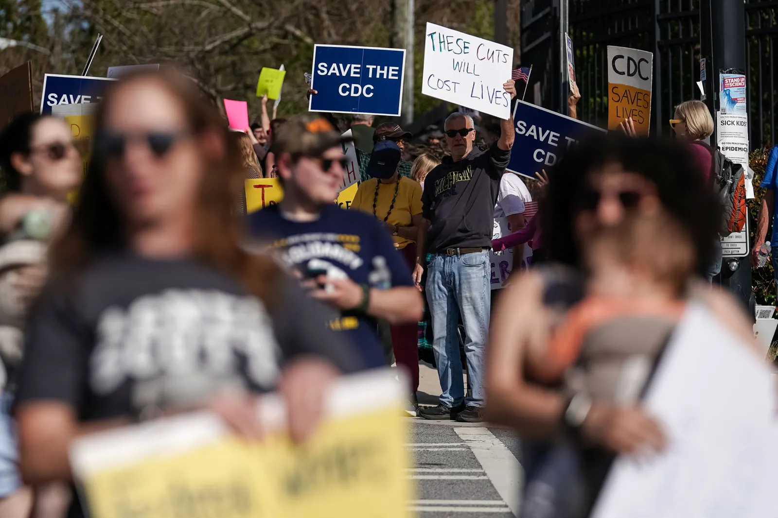 Blurred view of people standing in the foreground while a clear view of a man in jeans and a black sweater and sunglasses holds up a sign with one hand that reads ‘these cuts will cost lives’ while another sign to the left is blue with white words that read ‘save the CDC.’