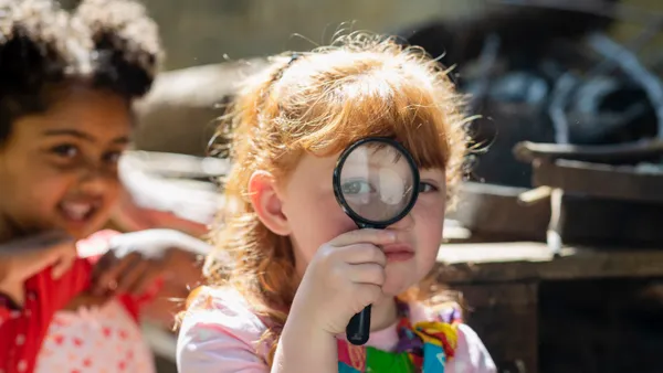 Young student looks through a magnifying glass toward the camera while another young student looks on.