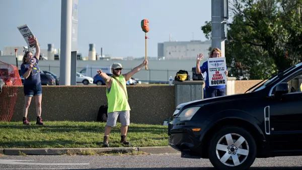 A black car passes by three people holding up signs that say on "On strike against Boeing."