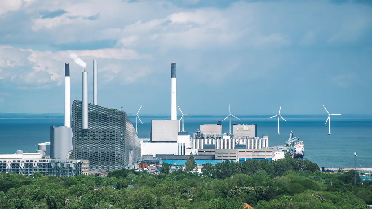 Industrial waste plant with smokestacks in Copenhagen, alongside waterfront with wind turbines in the background