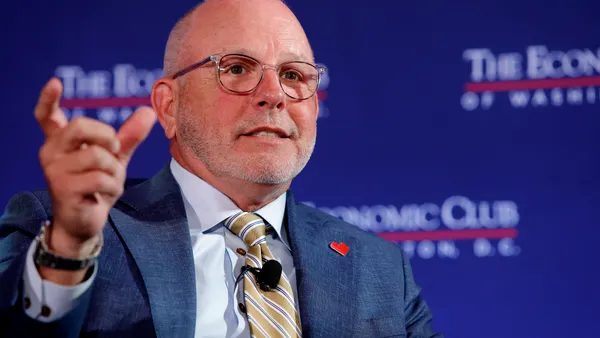 A seated man wearing glasses and a blue suit with a heart pin on his lapel gestures as he speaks.
