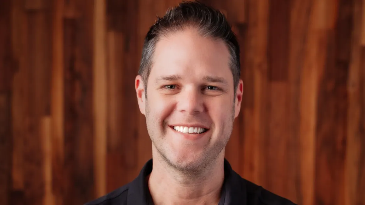 An image of a person wearing a black polo shirt smiling in front of a wood paneled background.