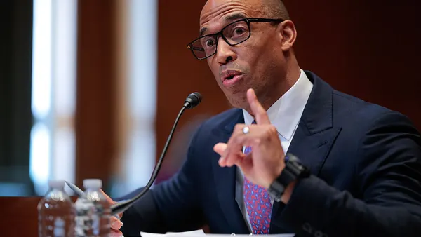 A person with glasses and in a suit and tie speaks into a microphone and gestures with his hand.