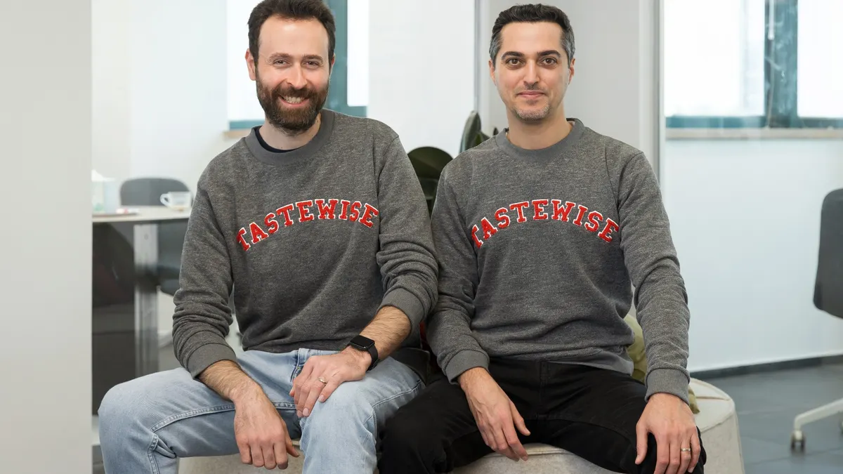 Tastewise founders Eyan Gaon and Alon Chen, wearing shirts with their company name, sit on a round bench in an office.