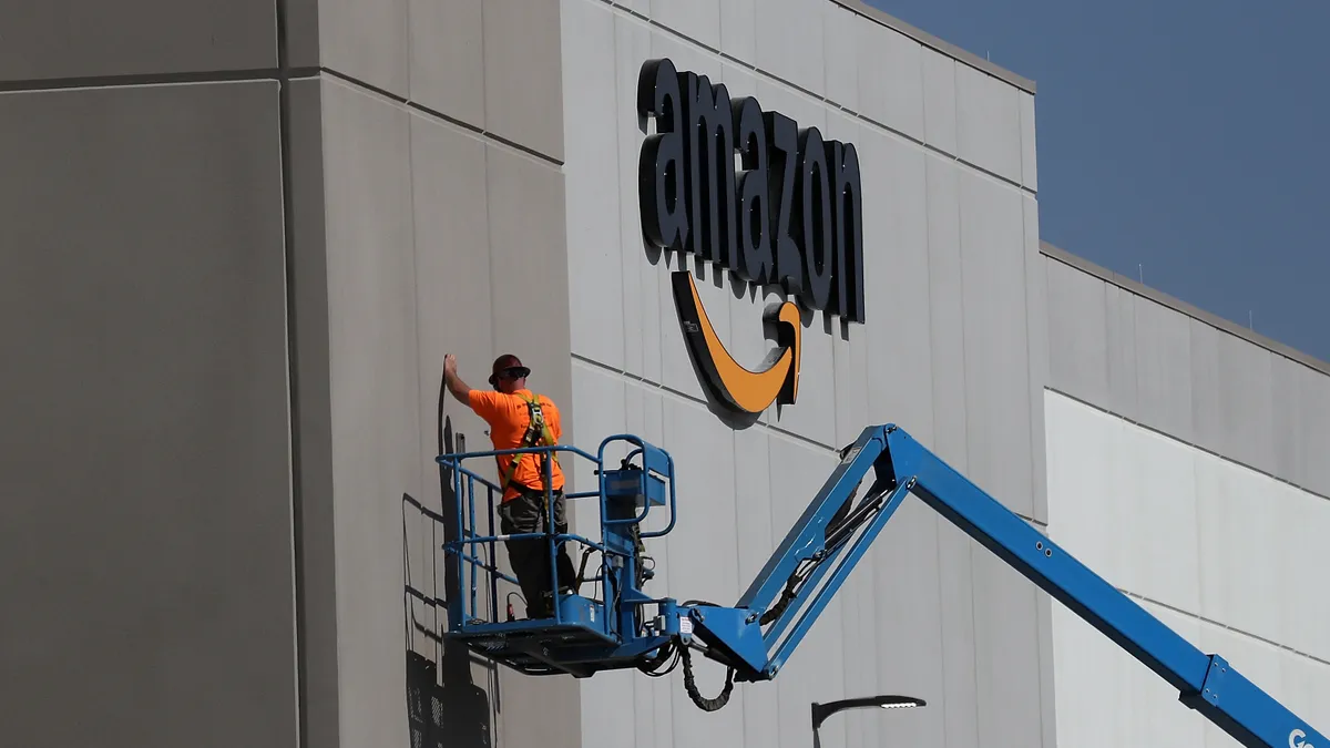 A worker on a blue crane makes repairs to the facade of a building housing an Amazon logo.