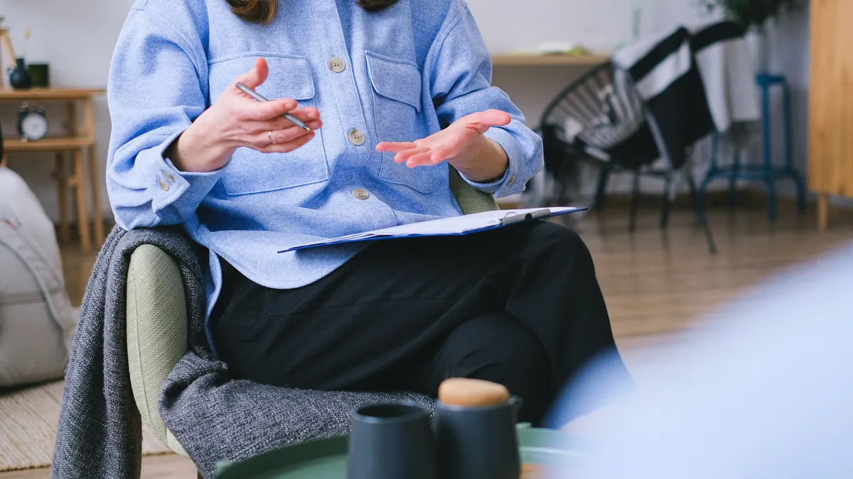 A faceless person, with a clipboard in the lap, speaks and gestures with their hands